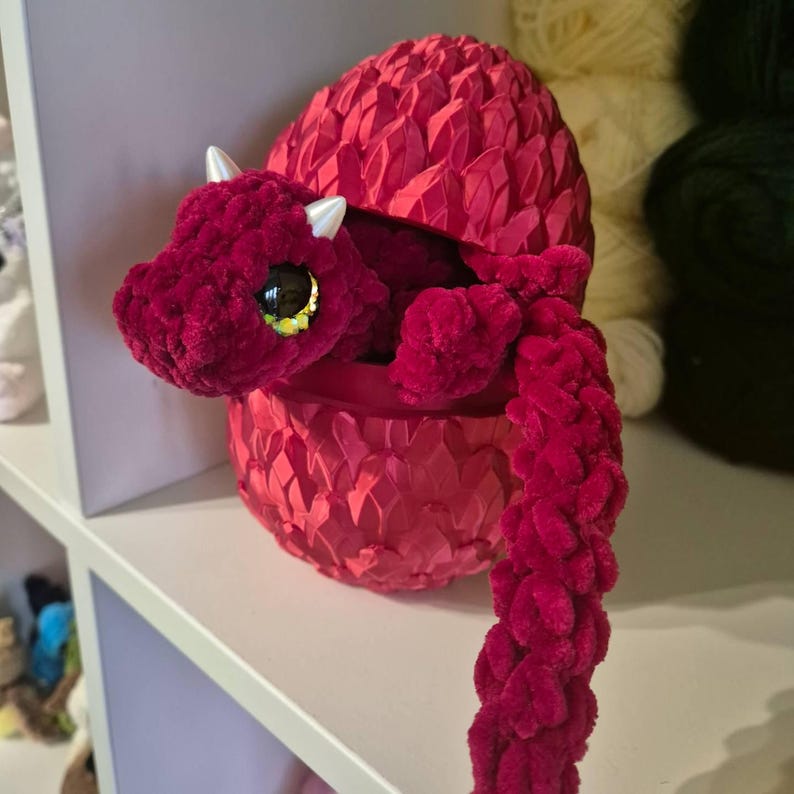 May include: A red crocheted dragon plush toy is inside a red crocheted egg-shaped container. The dragon has large, glittery eyes and a long tail.