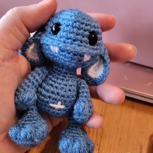 Op de afbeelding: Een klein blauw gehaakt monster met witte tanden en grote zwarte ogen. Het monster heeft kleine witte oren en wordt in een hand vastgehouden.