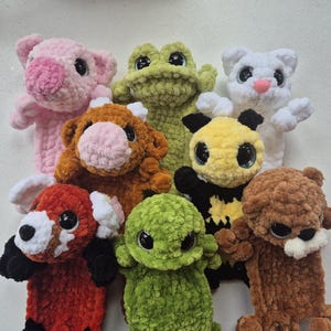 May include: A collection of handmade crochet stuffed animals in various colors. The assortment includes a pink pig, green frog, white cat, brown cow, red panda, green creature, yellow panda, and brown bear. Each has large, expressive eyes.