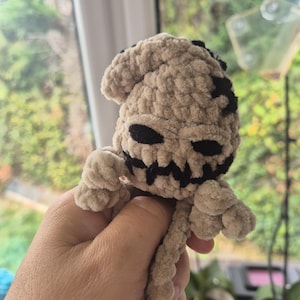 May include: A small, beige, plush crochet figure made of soft, fuzzy yarn. The figure features a rounded head with a stitched black face, including two oval eyes and a jagged, stitched mouth. It has small, rounded arms and a long, thin tail.