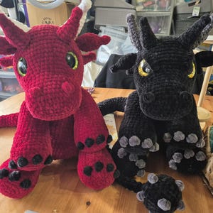 Peut inclure: Deux dragons au crochet faits à la main, un rouge et un noir, sont assis côte à côte. Le dragon rouge a des cornes à bout blanc et des yeux verts. Le dragon noir a des yeux jaunes. Les deux ont des griffes et des queues détaillées, sur le thème fantastique.