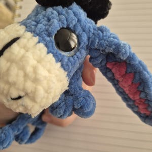 Peut inclure: Une peluche âne crochetée en fil bleu avec un museau crème, des cheveux en fil noir sur le dessus et de grands yeux en plastique noir. La longue oreille tombante présente une section intérieure rose, avec une texture douce et duveteuse.