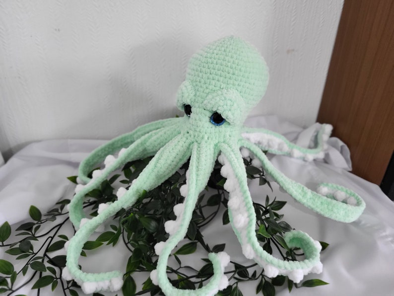 May include: A crocheted green octopus with white pom-poms on the ends of its tentacles. The octopus has blue eyes and is sitting on a bed of green leaves.
