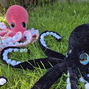May include: Two plush octopuses rest on green grass. One is pink with white and pink tentacles, the other is black with blue and white tentacles. Both have large, round eyes with a reflective quality.
