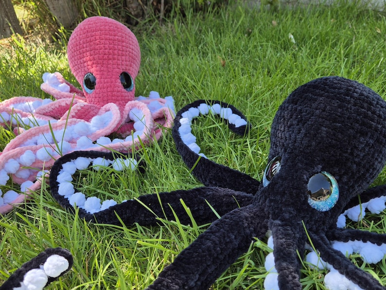 May include: Two plush octopuses rest on green grass. One is pink with white and pink tentacles, the other is black with blue and white tentacles. Both have large, round eyes with a reflective quality.