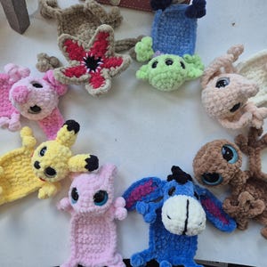 May include: A collection of ten handmade crochet plush bookmarks featuring various characters, including a red and beige flower-like creature, a yellow electric mouse, a blue donkey, a pink creature, and a brown alien, all with large plastic eyes.