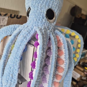 May include: A large, light blue crocheted octopus plush toy with large black eyes. The underside of the tentacles features colorful pom-poms in shades of purple, pink, yellow, and blue. The plush is soft and textured.
