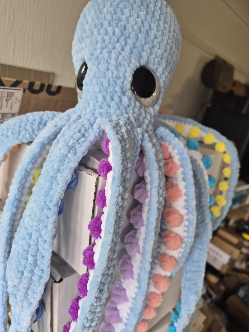 May include: A large, light blue crocheted octopus plush toy with large black eyes. The underside of the tentacles features colorful pom-poms in shades of purple, pink, yellow, and blue. The plush is soft and textured.