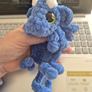 May include: A blue crocheted dragon plushie with white accents on its head and large, round, green and black eyes. The dragon has a curled tail and small, rounded feet. The plushie is made of a soft, textured yarn.