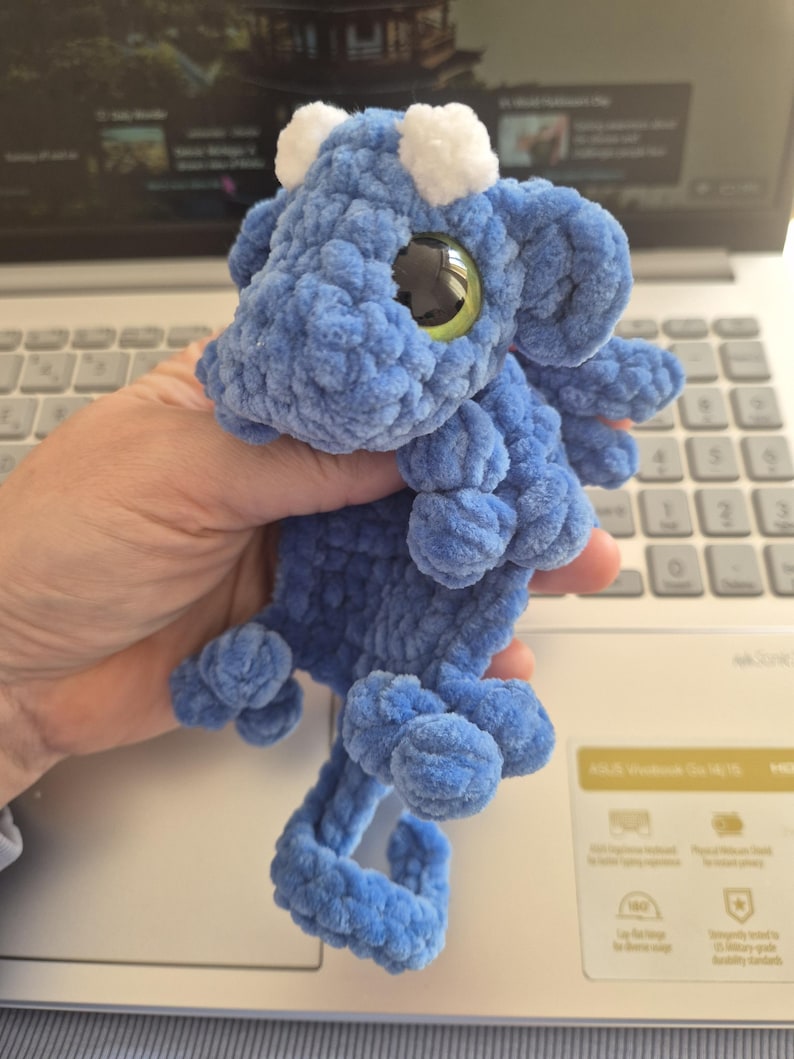 May include: A blue crocheted dragon plushie with white accents on its head and large, round, green and black eyes. The dragon has a curled tail and small, rounded feet. The plushie is made of a soft, textured yarn.