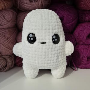 May include: White crocheted ghost plush toy with black eyes and a small black mouth. The toy is sitting on a background of pink and purple yarn balls.