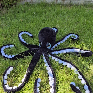 May include: A large, handmade black crochet octopus with white detailing on its tentacles. The octopus has large, expressive eyes with blue and white accents. It is lying on a bed of green grass, showcasing its soft, plush texture.