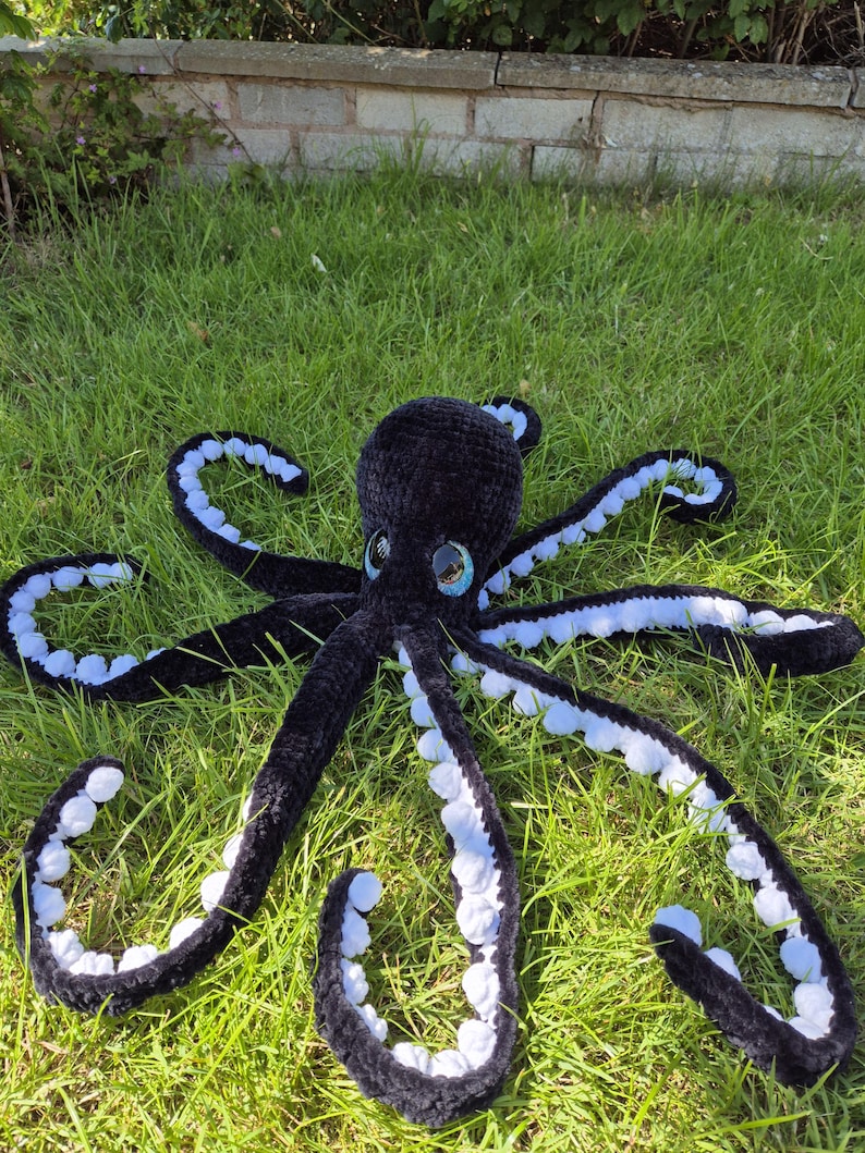 May include: A large, handmade black crochet octopus with white detailing on its tentacles. The octopus has large, expressive eyes with blue and white accents. It is lying on a bed of green grass, showcasing its soft, plush texture.