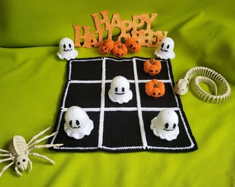 Puede incluir: Un tablero de juego de tres en raya tejido a crochet con fantasmas blancos y calabazas naranjas. El tablero es negro con líneas blancas y un borde blanco. El juego está configurado en una superficie verde con una araña esqueleto y una serpiente esqueleto.