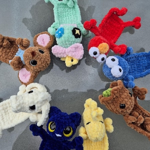 May include: A collection of eight crocheted plush bookmarks arranged in a circle. The characters include a brown mouse, a light green creature, a red monster, a blue monster, a brown tree-like figure, a yellow bird, a dark blue cat, and a white dog.