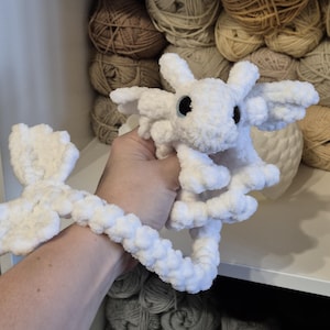 May include: White crocheted dragon plushie with a long tail. The dragon has blue eyes and is made with a fluffy yarn.