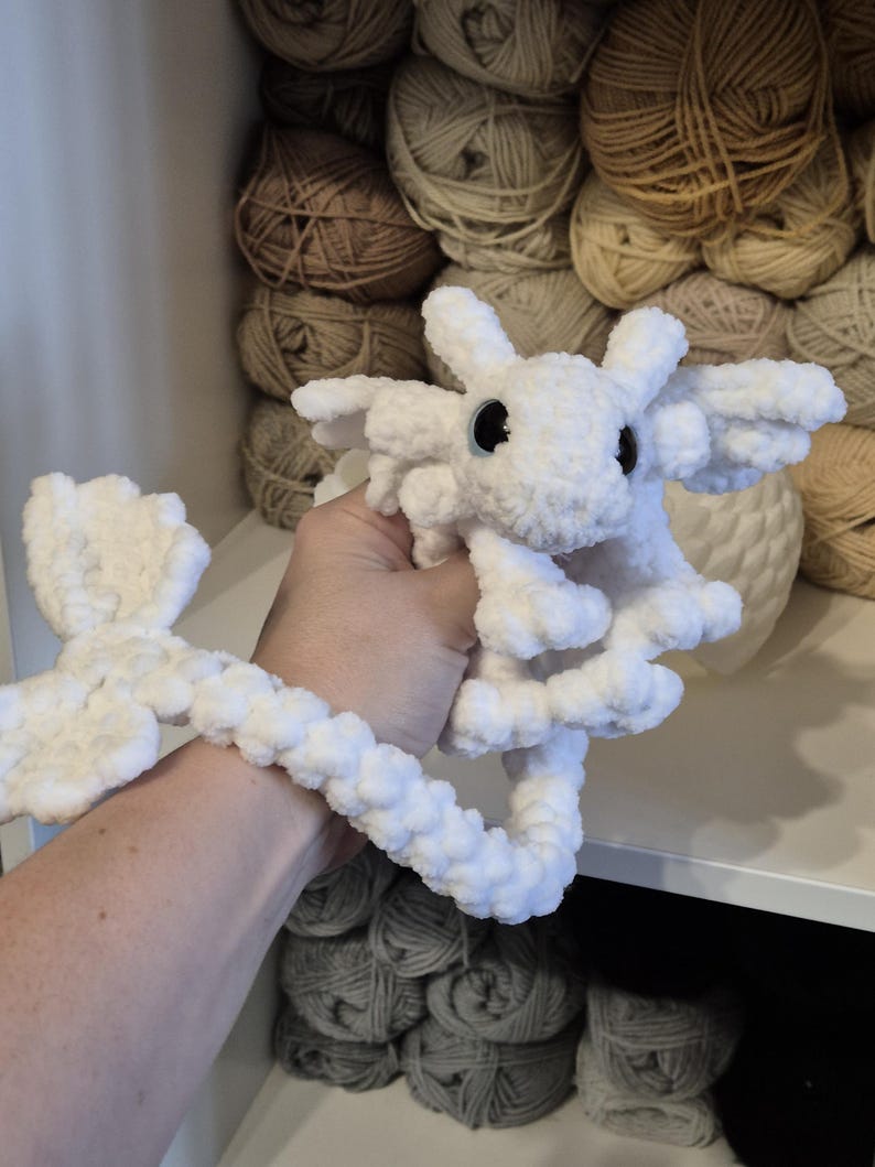 May include: White crocheted dragon plushie with a long tail. The dragon has blue eyes and is made with a fluffy yarn.