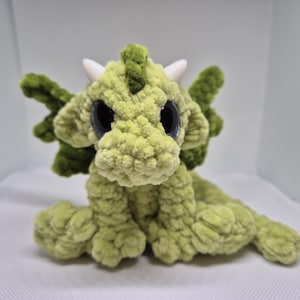 以下が含まれることがあります： 大きな黒い目と白い角を持つ、緑色の毛糸で編まれたドラゴンのぬいぐるみ。ドラゴンは白い表面に座っています。