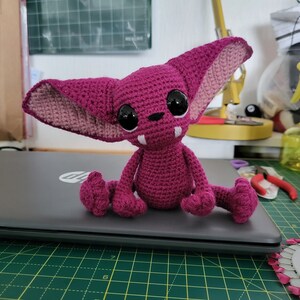 Puede incluir: Una criatura de crochet rosa con ojos grandes y colmillos. La criatura est&aacute; sentada en una computadora port&aacute;til.