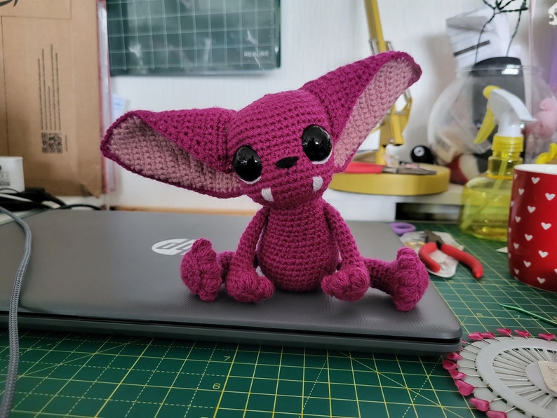 Puede incluir: Una criatura de crochet rosa con ojos grandes y colmillos. La criatura est&aacute; sentada en una computadora port&aacute;til.