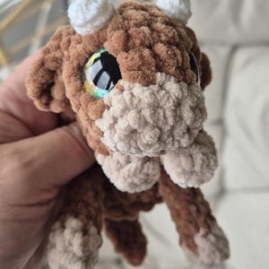 May include: A brown and beige crocheted stuffed animal with large, iridescent eyes. The animal has a fluffy texture and white crocheted horns.