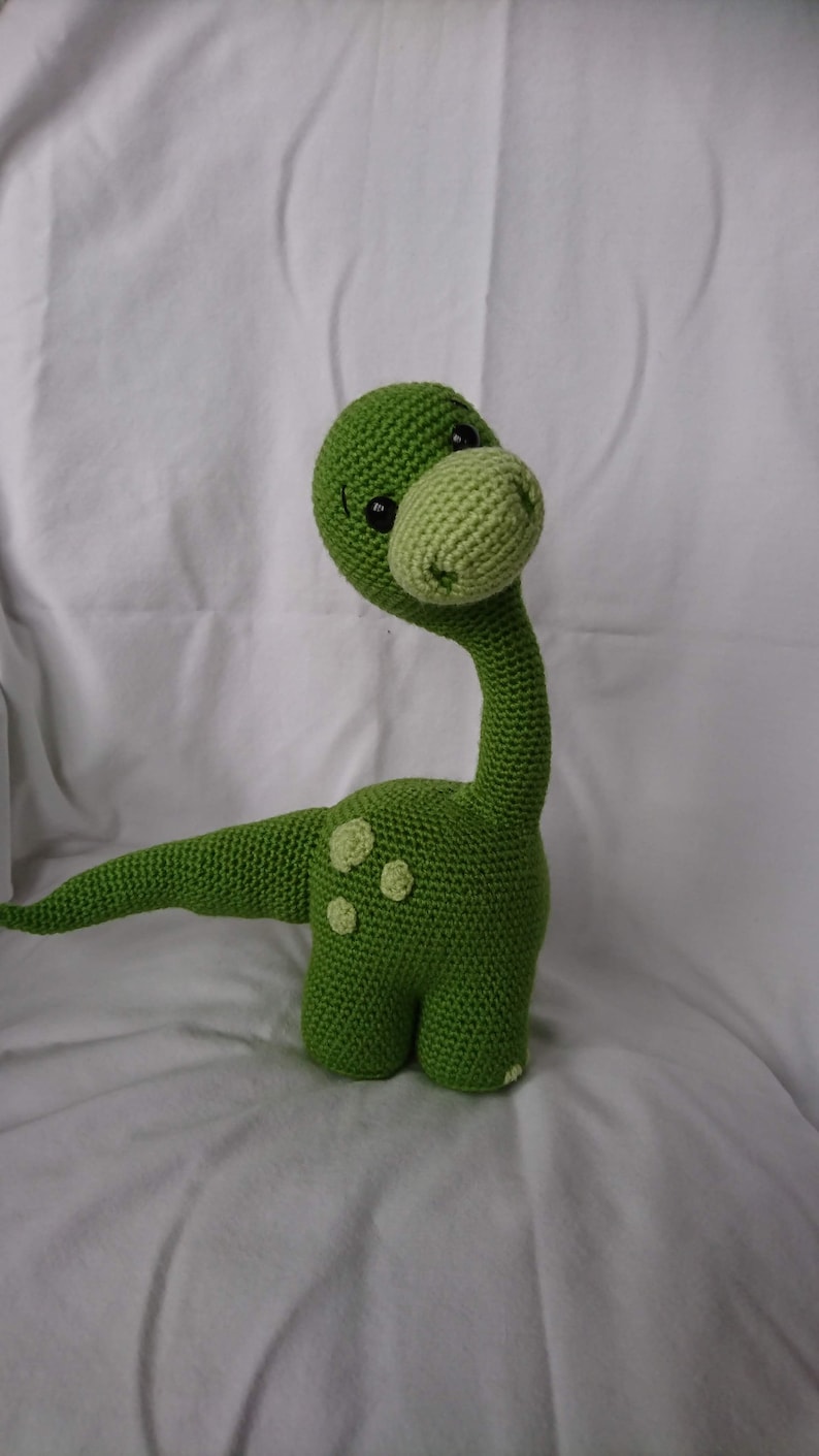 May include: A green crocheted dinosaur toy with black eyes and a long neck. The dinosaur has a light green nose and spots on its body.