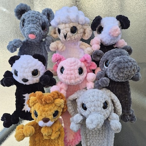 May include: A group of handmade crochet soft toys in different colours. The plush animals include a grey mouse, a white sheep, a black and white panda, a pink axolotl, a yellow lion, a grey elephant, a grey hippo, and a black and white cow.