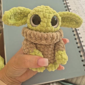 May include: A small, handmade crochet plush toy featuring a light green head with large black eyes and prominent ears, wearing a tan-colored textured sweater with light green accents on the sleeves and bottom, crafted from soft, chunky yarn.