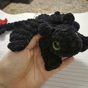 May include: A handmade black crochet dragon with green eyes and a red fin. The plush toy is held in a hand and is made of a soft, textured yarn. The dragon has a detailed, rounded shape.