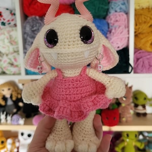 May include: A crocheted plush toy with pink horns, a pink dress, and sparkly purple eyes. The toy is holding a pink heart in its hand.