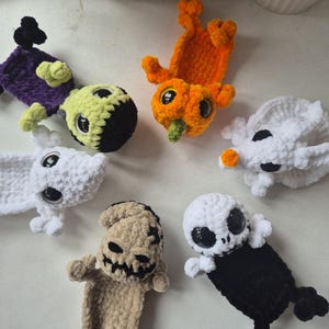 May include: A collection of six crocheted plush figures featuring a green-faced character with purple clothing, an orange pumpkin-like figure, two white ghost-like creatures, a tan burlap-textured character, and a white skull-faced figure with black clothing.