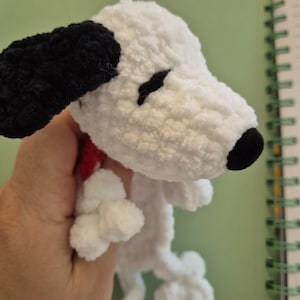 May include: A small, white, crocheted plush dog with a black ear, a black nose, and black embroidered eyes. The plush figure features a red collar around its neck and is crafted from a soft, textured yarn, showcasing a handmade aesthetic.