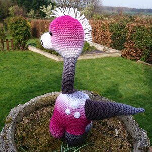 May include: A pink and white crocheted dinosaur toy with a white spiky crest. The dinosaur is sitting in a stone planter with green moss.