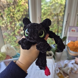 May include: A black crocheted plush toy resembling a dragon with large, bright green eyes and a small red accent on the tip of its tail. The creature features a soft, textured yarn finish, rounded ears, and small wings extending from its sides.