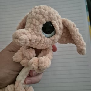 May include: A light beige crochet plush toy featuring a textured, soft yarn finish, large black plastic eyes with a subtle blue rim, long floppy ears, and a small snout. The creature has a rounded head and a slender body with small limbs.