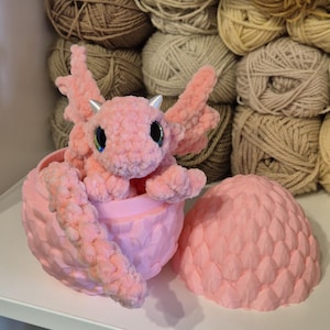 May include: A pink crocheted dragon figurine sitting inside a pink dragon egg. The egg is open and the dragon is peeking out.