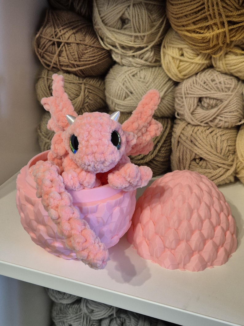 May include: A pink crocheted dragon figurine sitting inside a pink dragon egg. The egg is open and the dragon is peeking out.