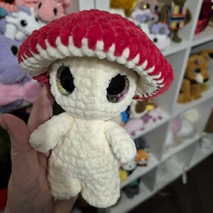 May include: A crocheted mushroom plush toy with a red and white cap and white body. The toy has large, glittery eyes.