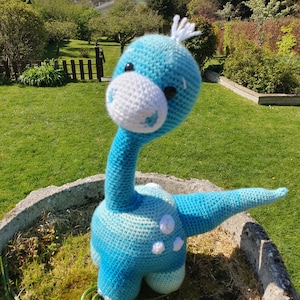 May include: A blue and white crocheted dinosaur toy with white spots. The dinosaur is standing on a bed of green grass.