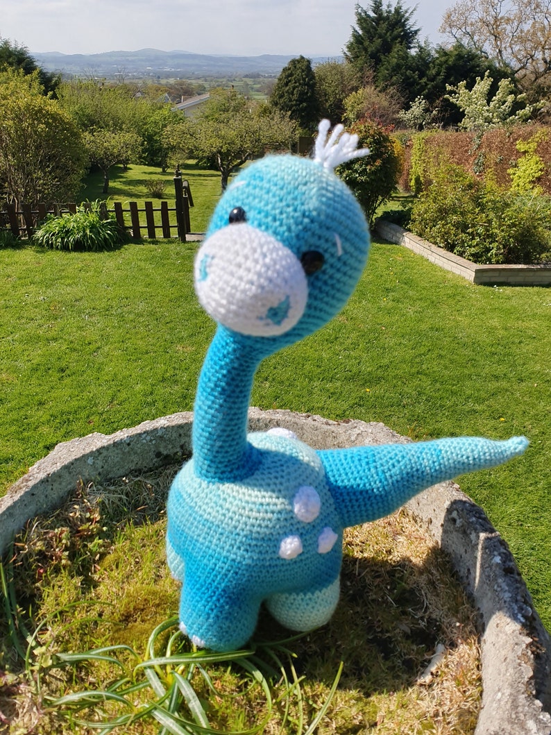 May include: A blue and white crocheted dinosaur toy with white spots. The dinosaur is standing on a bed of green grass.