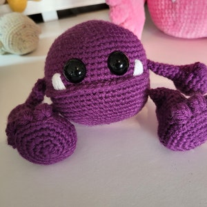 May include: A purple crocheted monster with large black eyes and white teeth. The monster has three arms, each with a rounded end.