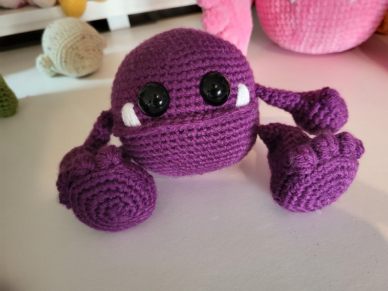 May include: A purple crocheted monster with large black eyes and white teeth. The monster has three arms, each with a rounded end.