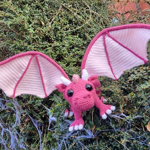 May include: A pink crocheted dragon with large eyes and wings. The dragon is sitting on a bed of green leaves.