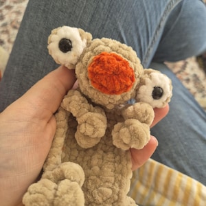 May include: A beige crochet plush toy featuring a textured, soft yarn body with a prominent orange circular nose and two large, white, protruding eyes with black pupils. The creature has small, rounded limbs and a flat, elongated body shape.