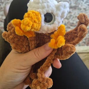 May include: A crocheted plush eagle toy featuring a white head, brown wings and body, and bright orange feet and beak. The eagle has large, round, black plastic safety eyes and is crafted from a soft, textured yarn material with a plush finish.