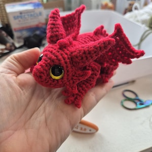 May include: A small, red crocheted dragon toy with large, yellow and black eyes, wings, and horns. The dragon is handmade with detailed crochet work. The image is a close-up shot of the dragon, highlighting its whimsical design and features.