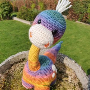 May include: A rainbow crocheted dinosaur toy with white yarn for the horns and eyes. The dinosaur is sitting on a bed of green moss.