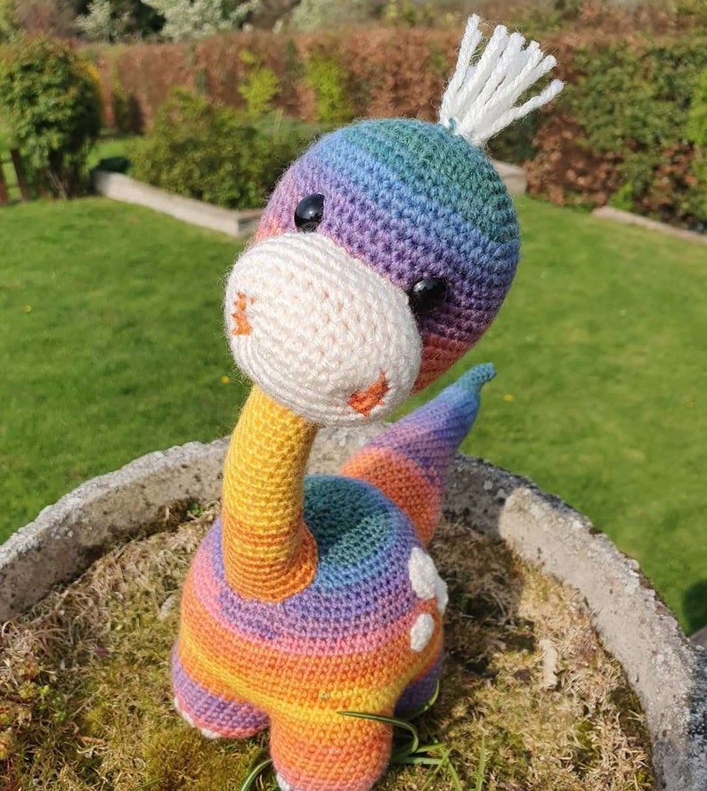 May include: A rainbow crocheted dinosaur toy with white yarn for the horns and eyes. The dinosaur is sitting on a bed of green moss.