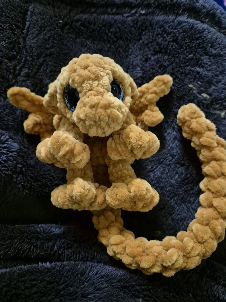 May include: A crocheted brown dragon with blue eyes. The dragon has wings and a long tail.