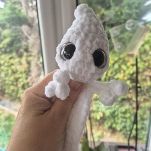May include: A white, plush, crocheted creature with a teardrop-shaped head and a long, slender body. It features two large, round, black plastic eyes with a glossy finish and small, textured, multi-fingered appendages extending from the sides.